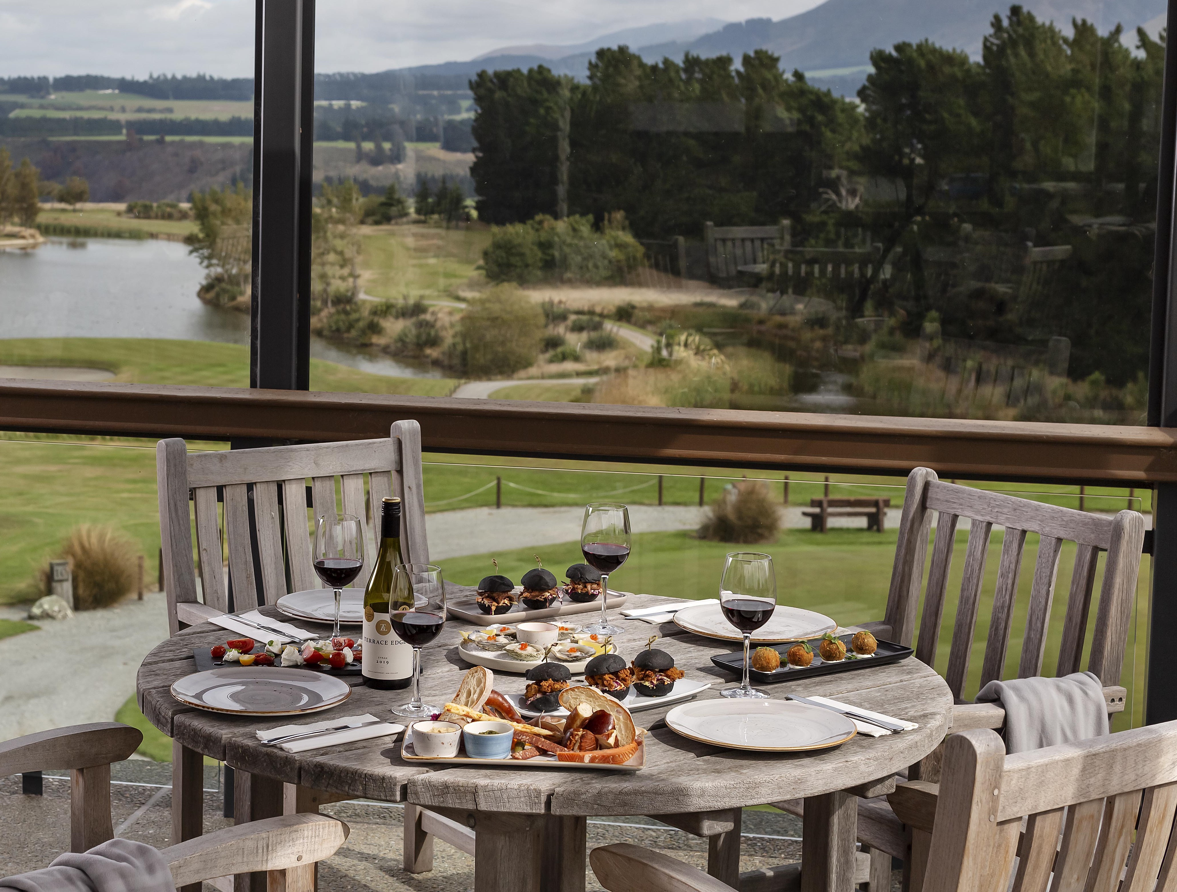 Outdoor dining table overlooking a golf course and lake, featuring wine bottles, glasses of red wine, sliders, oysters and platters.